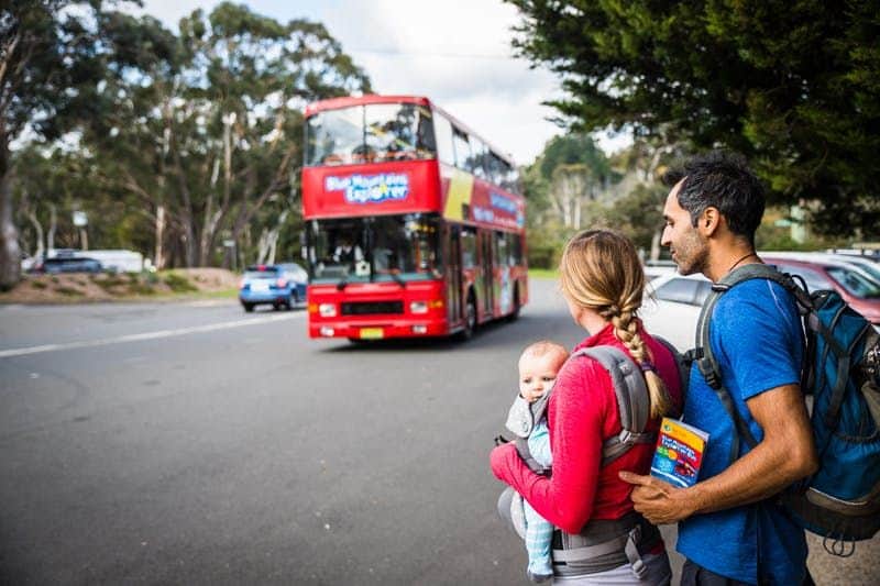 Blue Mountains Sydney: Hop-on Hop-off Bus + Scenic World Unlimited Rides Pass A couple with a baby in a carrier stand in a parking lot, next to parked cars, while a red double-decker bus approaches.