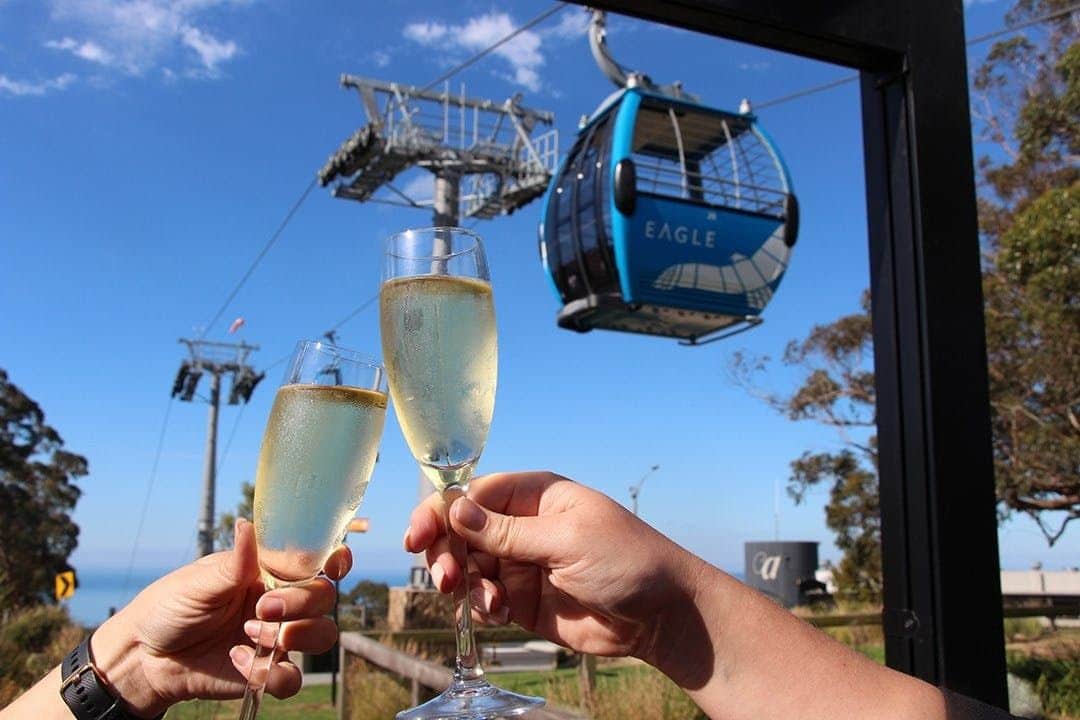 Arthurs Seat Eagle: Roundtrip Gondola Ticket + Drink Two people clinking champagne glasses with a blue cable car labeled "Eagle" against a clear sky background.
