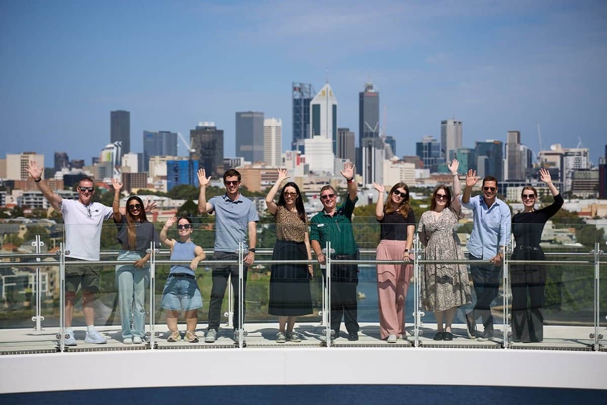 A group of people wave while standing on a bridge with a city skyline in the background on a sunny day.