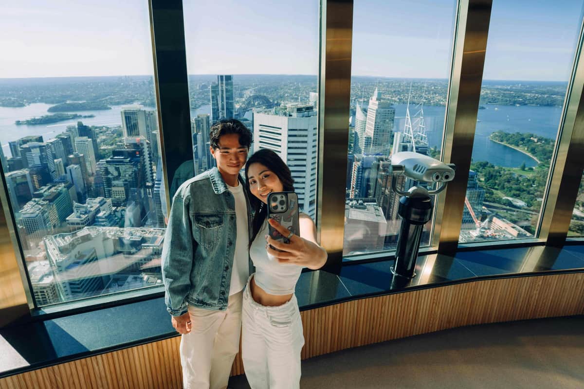 Sydney Tower Eye: Entry Ticket Two people take a selfie in a skyscraper with large windows, overlooking a cityscape with tall buildings and distant water views.