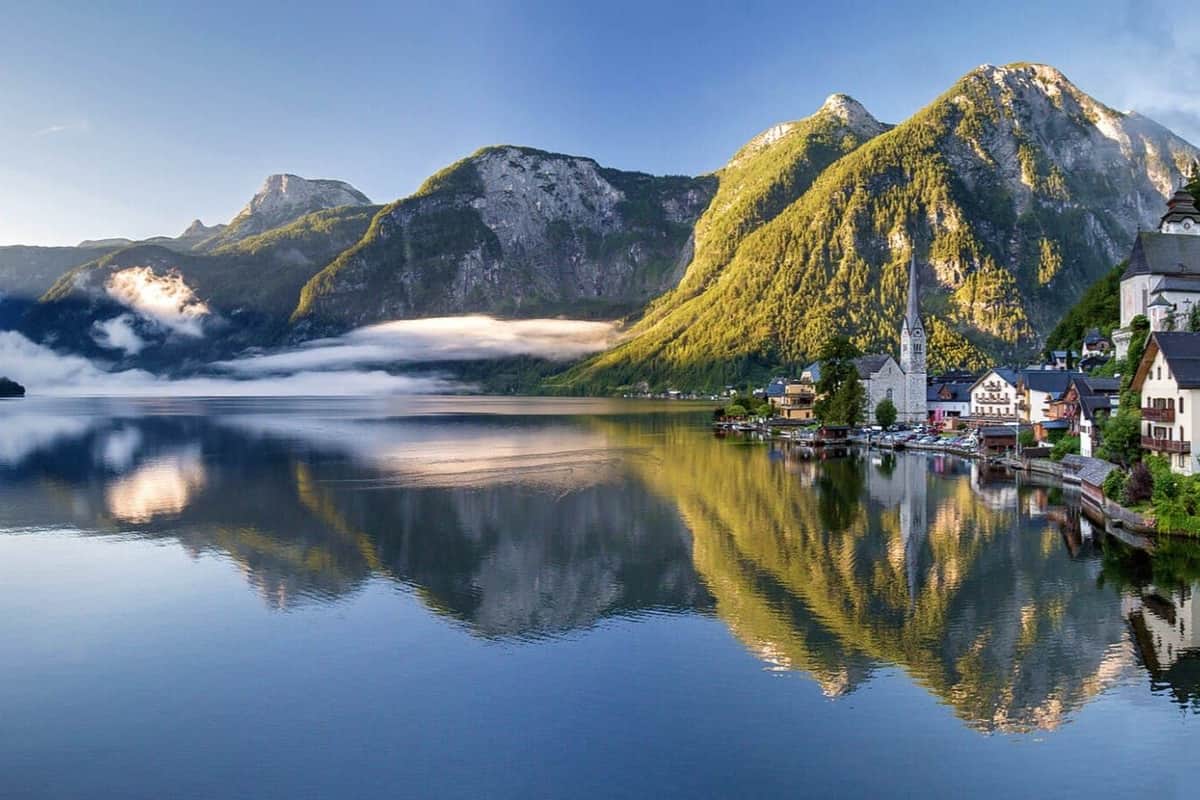 A lakeside village with a church spire, surrounded by green mountains reflecting in calm water under a clear blue sky.