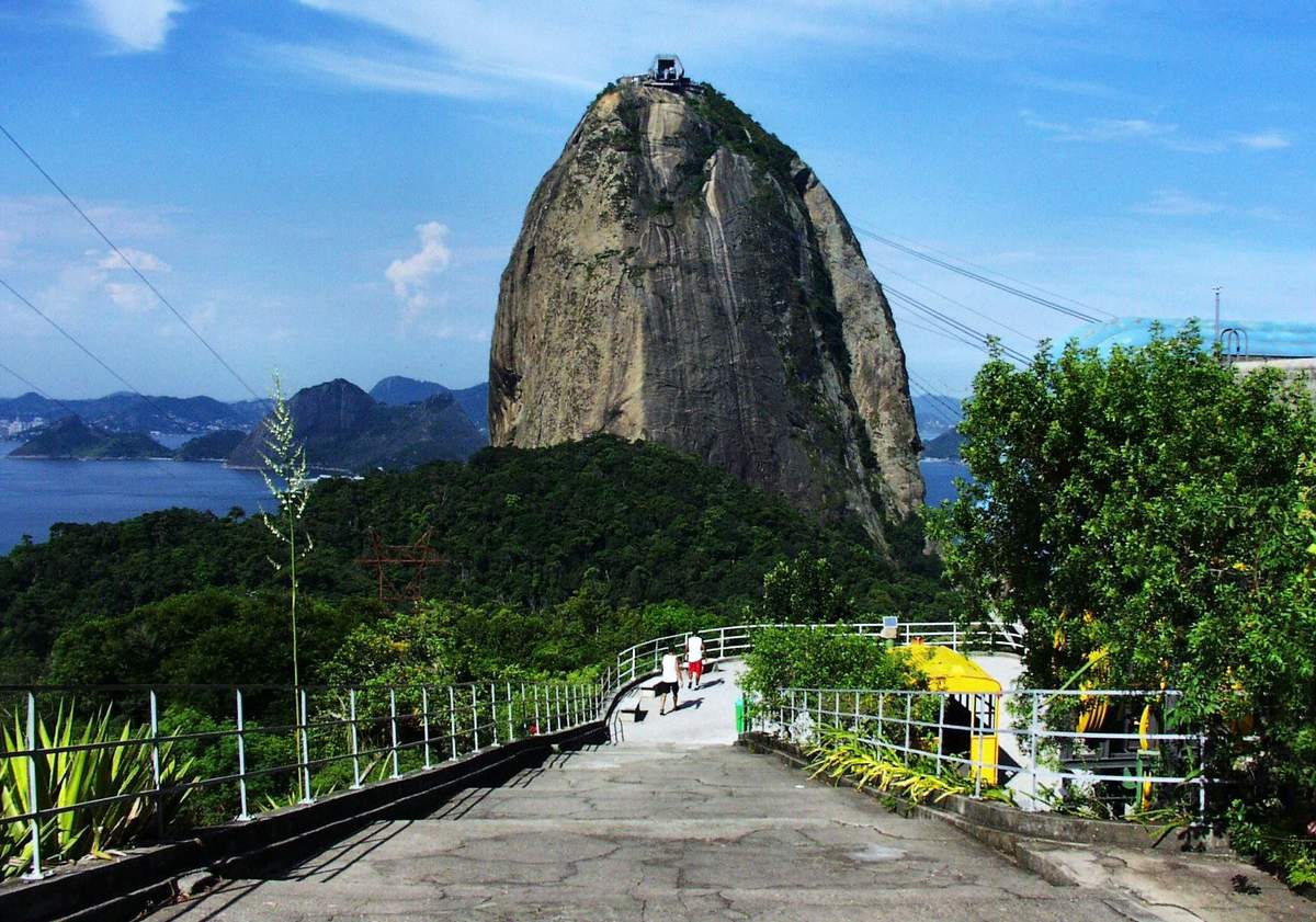Sugarloaf City Tour & Cable Car Ride A steep rock formation surrounded by greenery under a blue sky with a paved pathway and railing in the foreground.