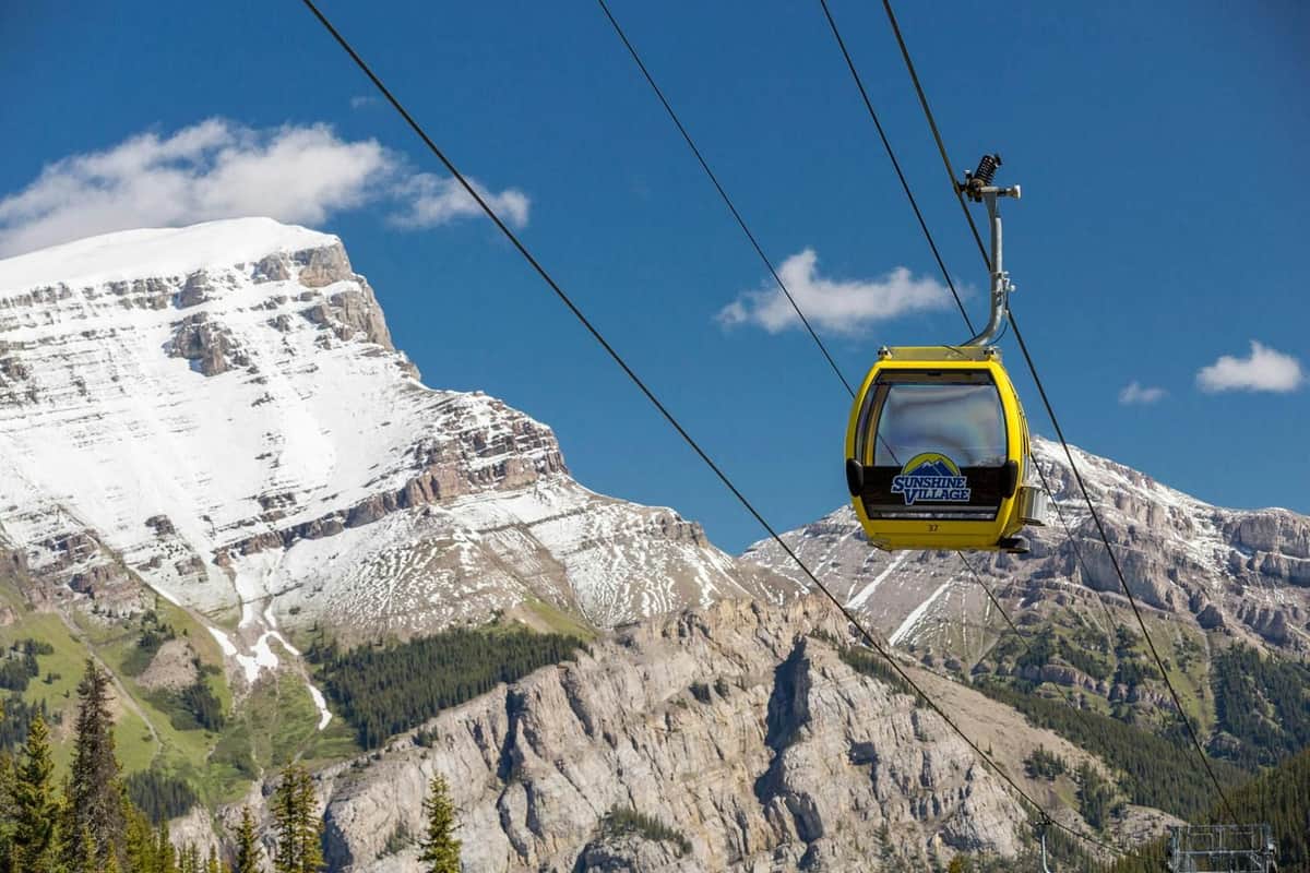 Banff Sunshine Village: Entry Ticket + Gondola & Chairlift Access A yellow gondola labeled "Sunshine Village" rides on cables against a backdrop of snow-capped mountains and blue sky.