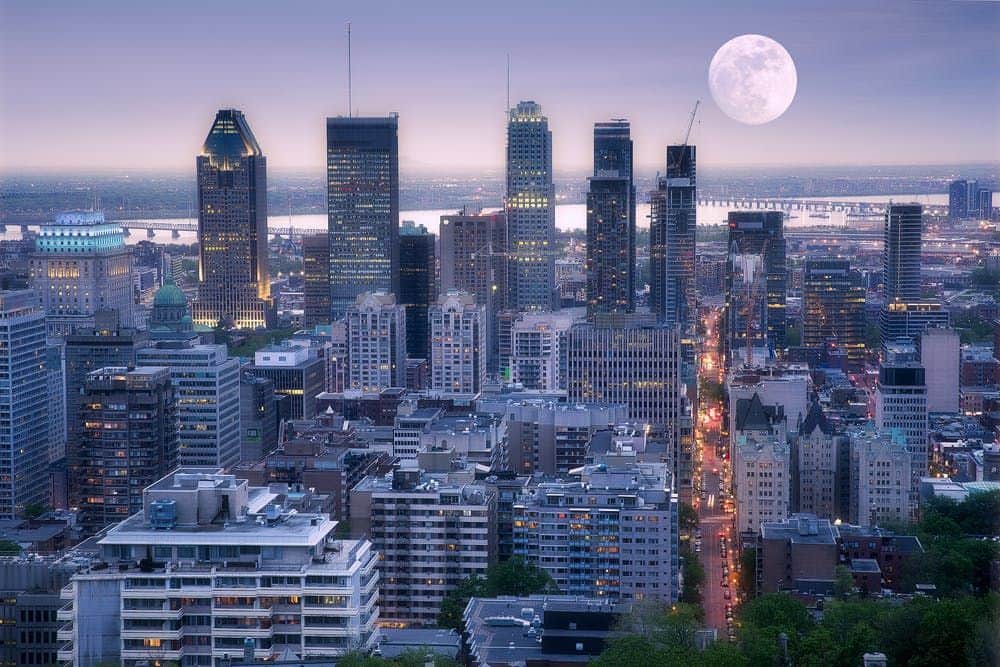 Montreal: Scenic Night Tour + La Grande Roue de Montreal Entry Ticket City skyline at dusk with tall buildings and a full moon rising. Lights illuminate the streets and buildings.