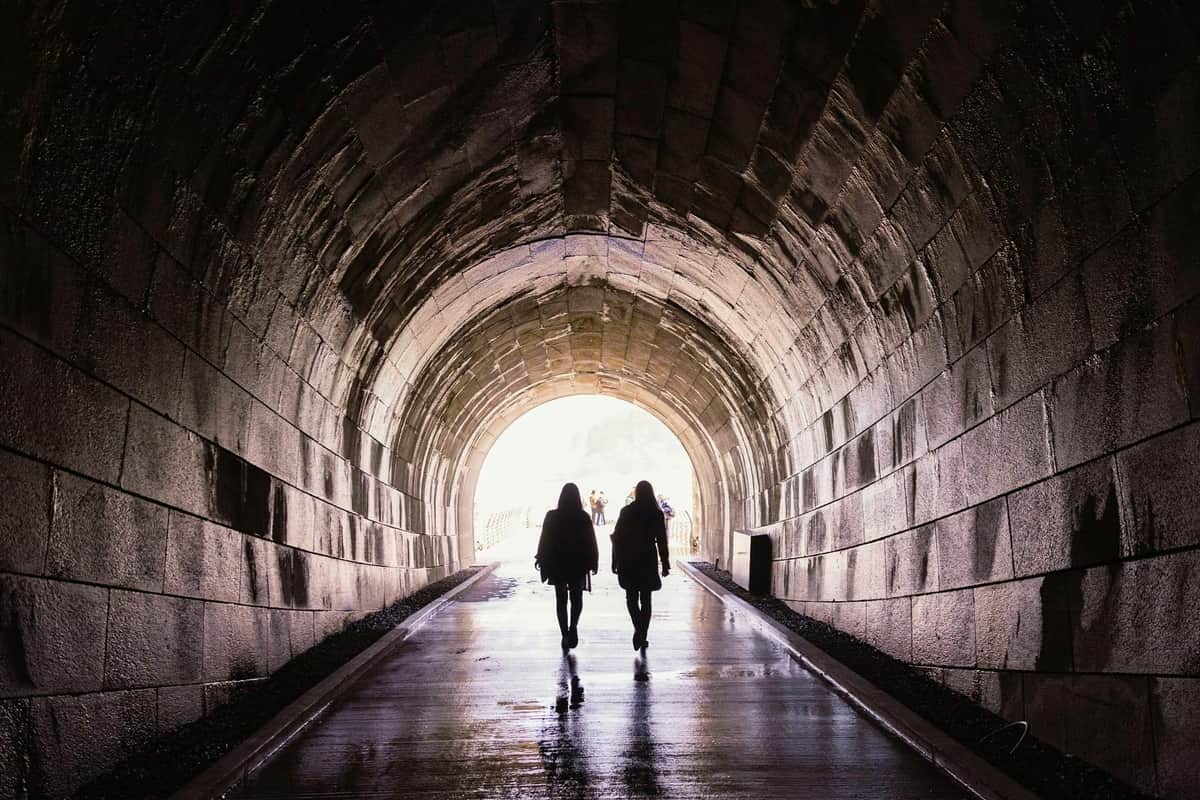 Niagara Falls Power Station Tour with Skylon Tower & Tailrace Tunnel Experience Two people walk through a stone archway tunnel, heading towards a bright light at the end. The ground is wet and reflective.