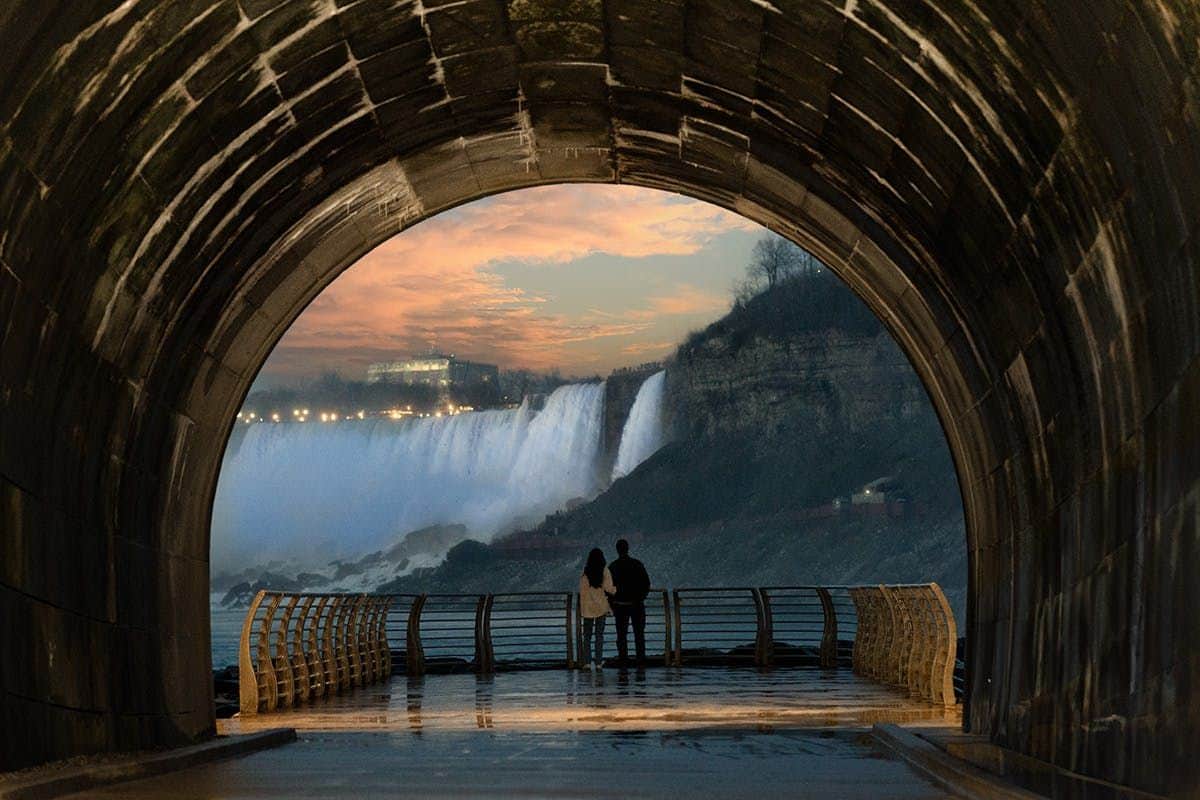 The Tunnel at The Niagara Parks Power Station: Evening Entry + Light Show Two people stand in a tunnel, looking out at a large waterfall against a sunset-lit sky.