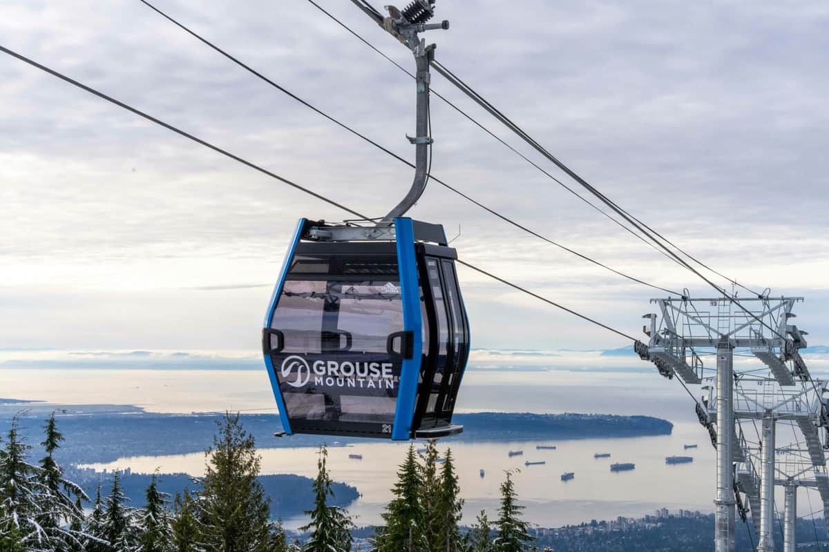 A Grouse Mountain cable car suspended above a scenic landscape with trees, water, and distant ships and islands.