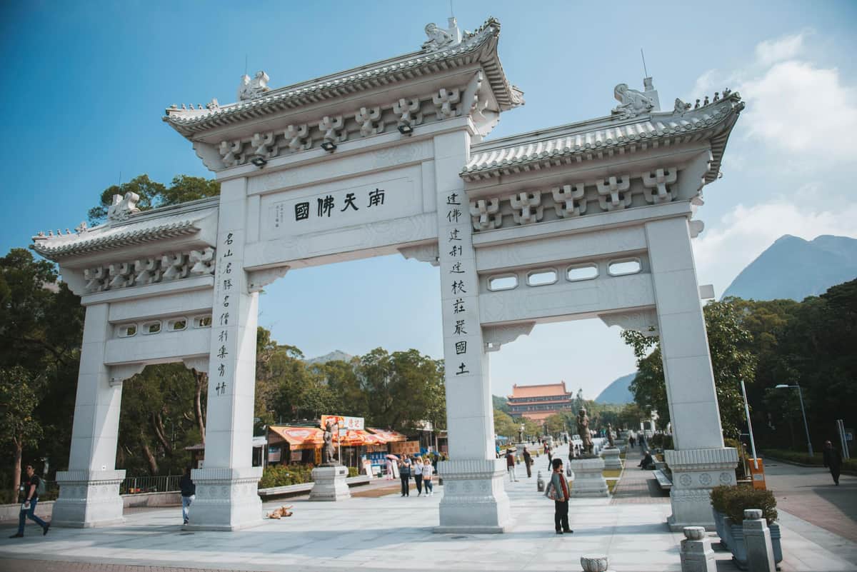 360 Lantau Culture & Heritage Insight Tour + Cable Car White traditional Chinese archway with ornate designs. People walking and trees in the background under a clear blue sky.