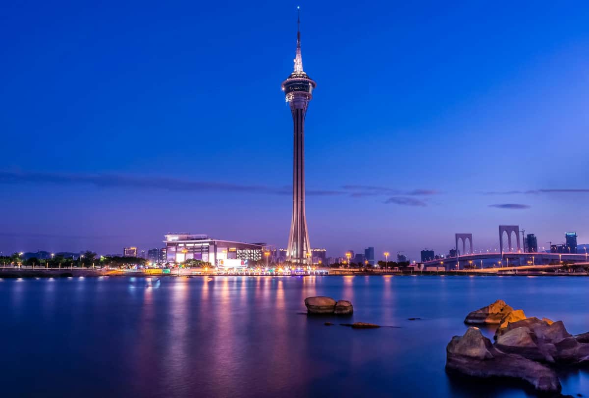 Macau Tower A tall tower with observation deck and antennas stands by a waterfront, illuminated city lights reflecting on the water at dusk.