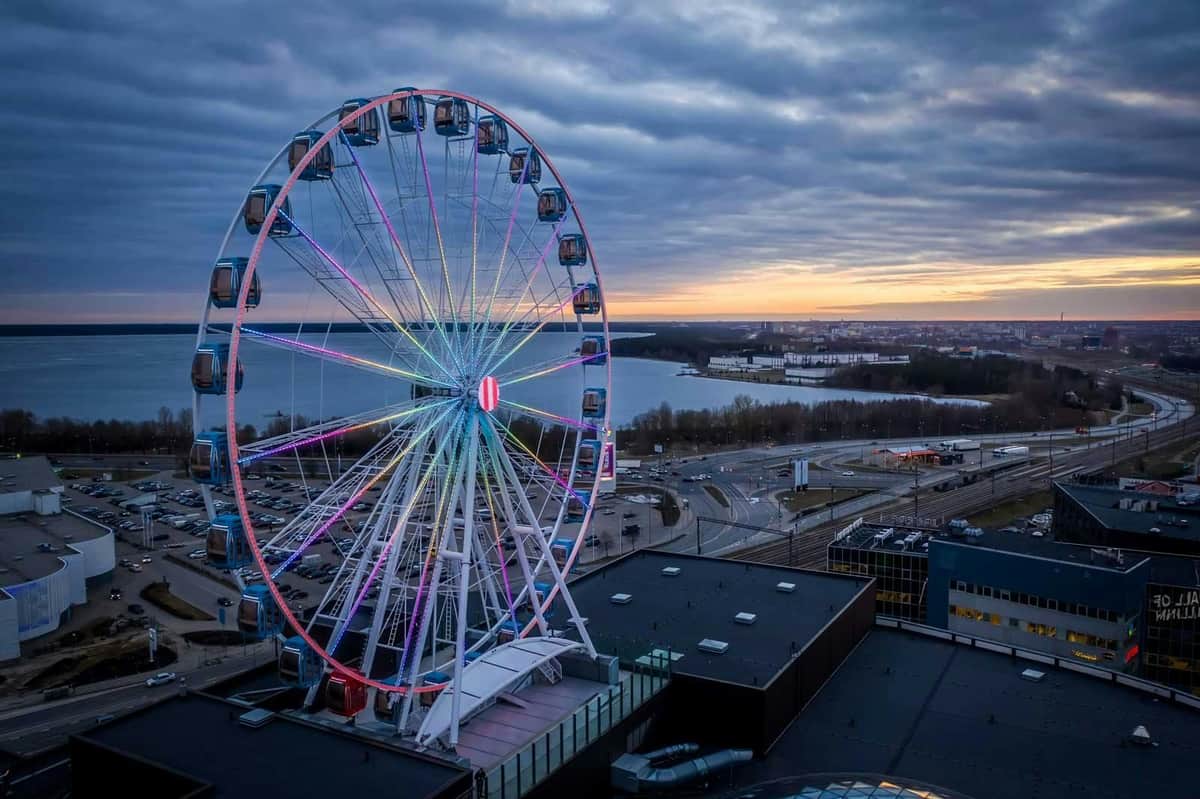 Skywheel of Tallinn: Entry Ticket A Ferris wheel with colorful lights stands near a body of water at sunset, with buildings and roads visible below.