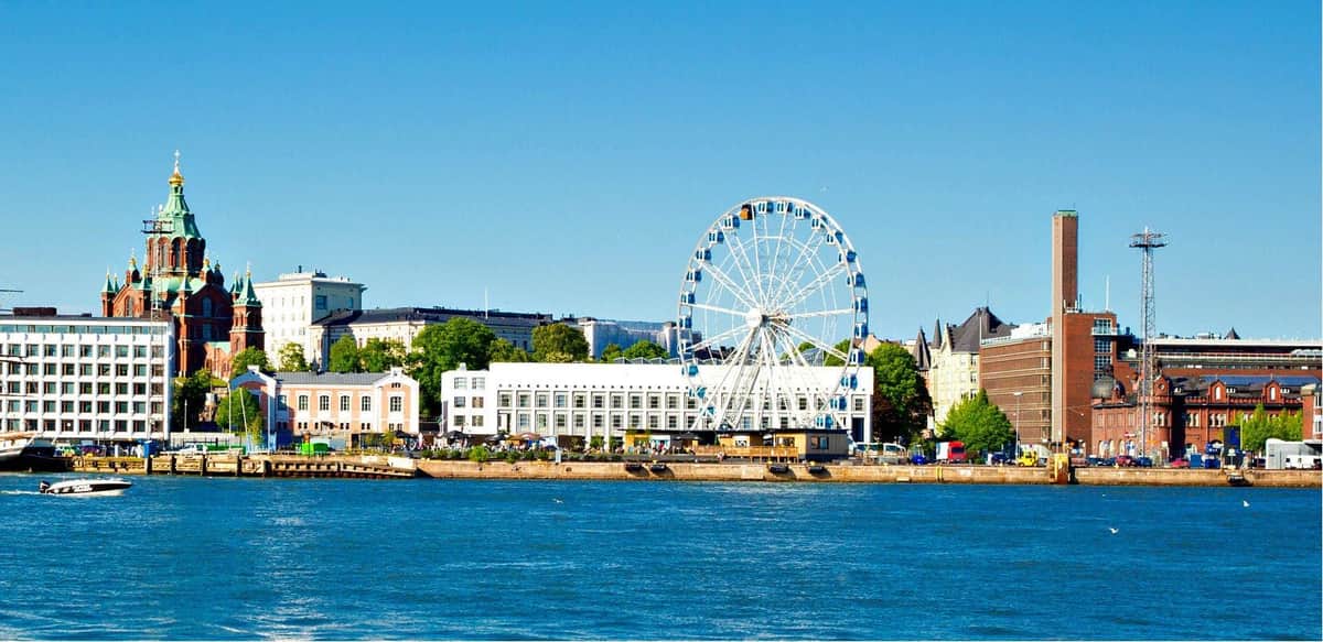 SkyWheel Helsinki: Entry Ticket A large Ferris wheel beside a waterfront, with buildings and trees in the background under a clear blue sky.