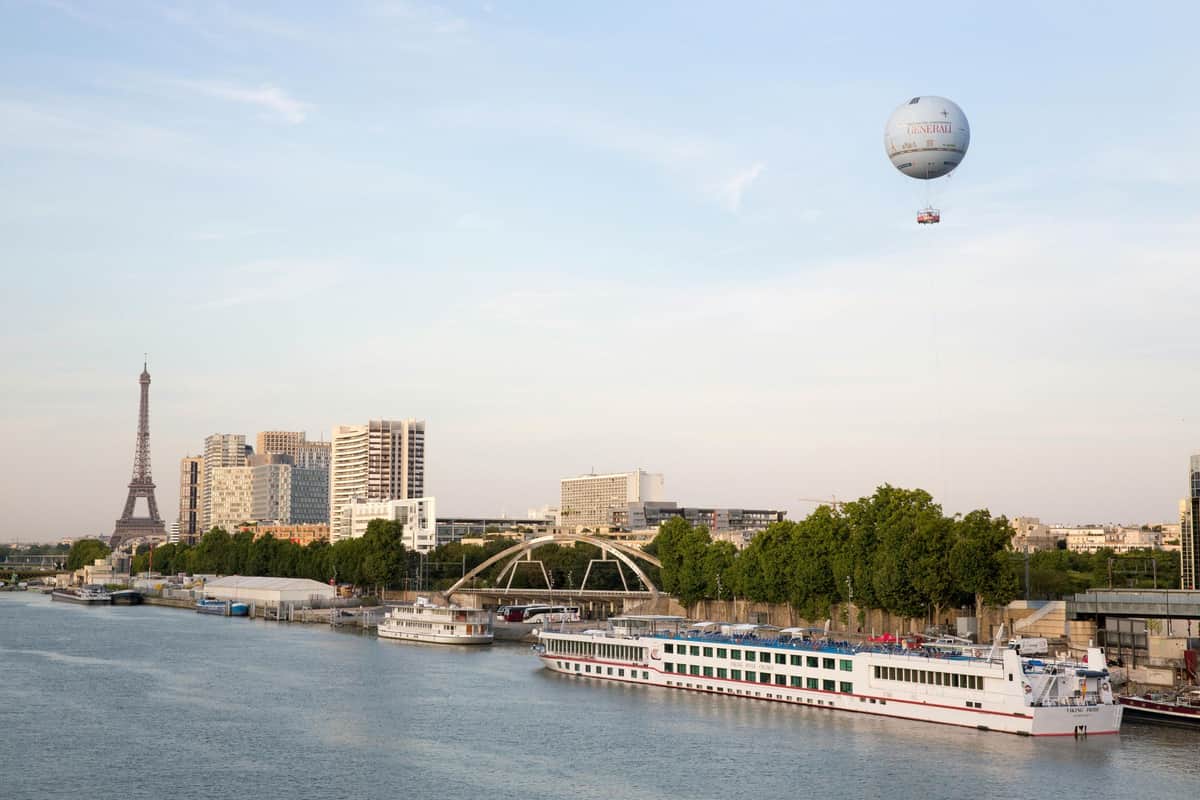 Ballon de Paris GENERALI & Bateaux Mouches Seine River Cruise A riverside scene with docked boats, buildings, a bridge, trees, and a tethered hot air balloon in the sky.