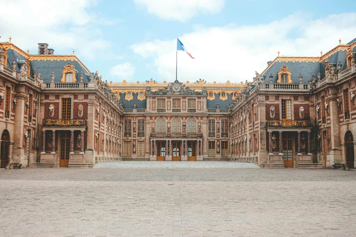 Eiffel Tower & Versailles Palace: Day Tour from Paris + Roundtrip Train Tickets A grand palace with ornate architecture, gold accents, and a French flag atop, viewed from a cobblestone courtyard.