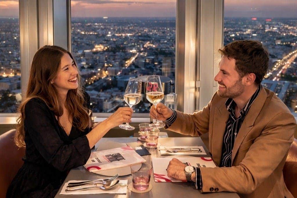 Berlin TV Tower: Admission + Four Course My Home - Tim Raue Menu + Drinks Two people clinking wine glasses at a restaurant table during sunset, with a cityscape visible through the window.
