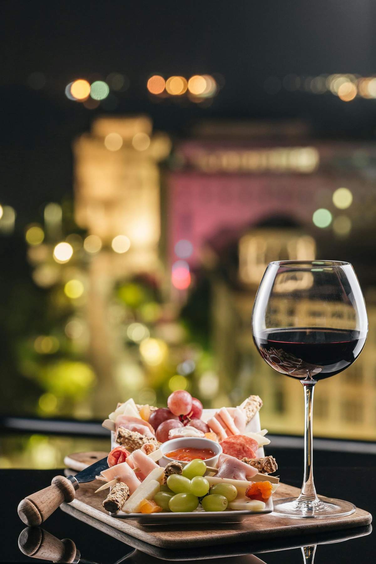 A glass of red wine and a charcuterie board with meats, cheeses, grapes, and crackers in front of a blurred cityscape at night.