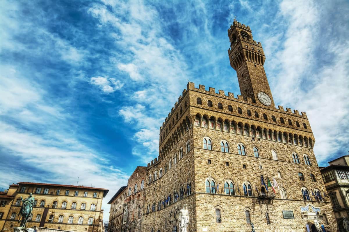 Palazzo Vecchio, Arnolfo Tower, Museum: Entry Ticket + Audio Guide Historic stone building with arched windows and a tall clock tower under a partly cloudy blue sky. Statues line the building's facade.