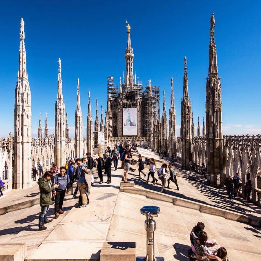 Duomo di Milano: Entry Ticket + Rooftop Access People walking and sightseeing on the rooftop terrace of a Gothic cathedral with intricate spires and clear blue sky.