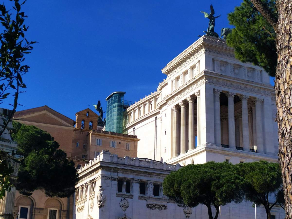 Altar of the Fatherland: Glass Elevator Skip The Line Ticket + Audio App Historic buildings with columns and statues against a blue sky, partially obscured by trees in the foreground.