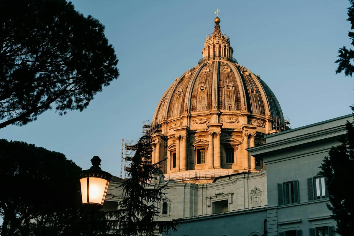 St. Peter's Basilica: Guided Tour + Dome Access A large, ornate dome with scaffolding, surrounded by trees and a lit streetlamp, set against a clear sky.