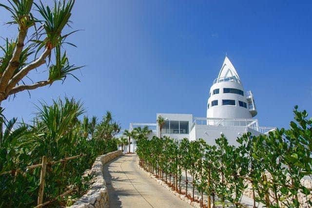 Kouri Ocean Tower A white tower with a unique pointed roof stands amid lush greenery along a winding pathway under a clear blue sky.