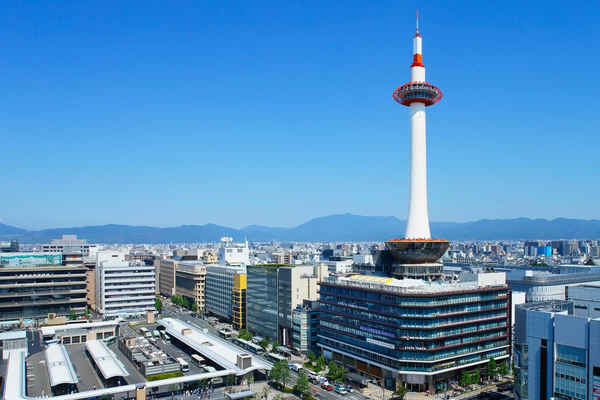 Nidec Kyoto Tower : Billet d'entrée Une haute tour d'observation au sommet orange et blanc se dresse parmi des bâtiments urbains dans un ciel bleu clair avec des montagnes au loin.