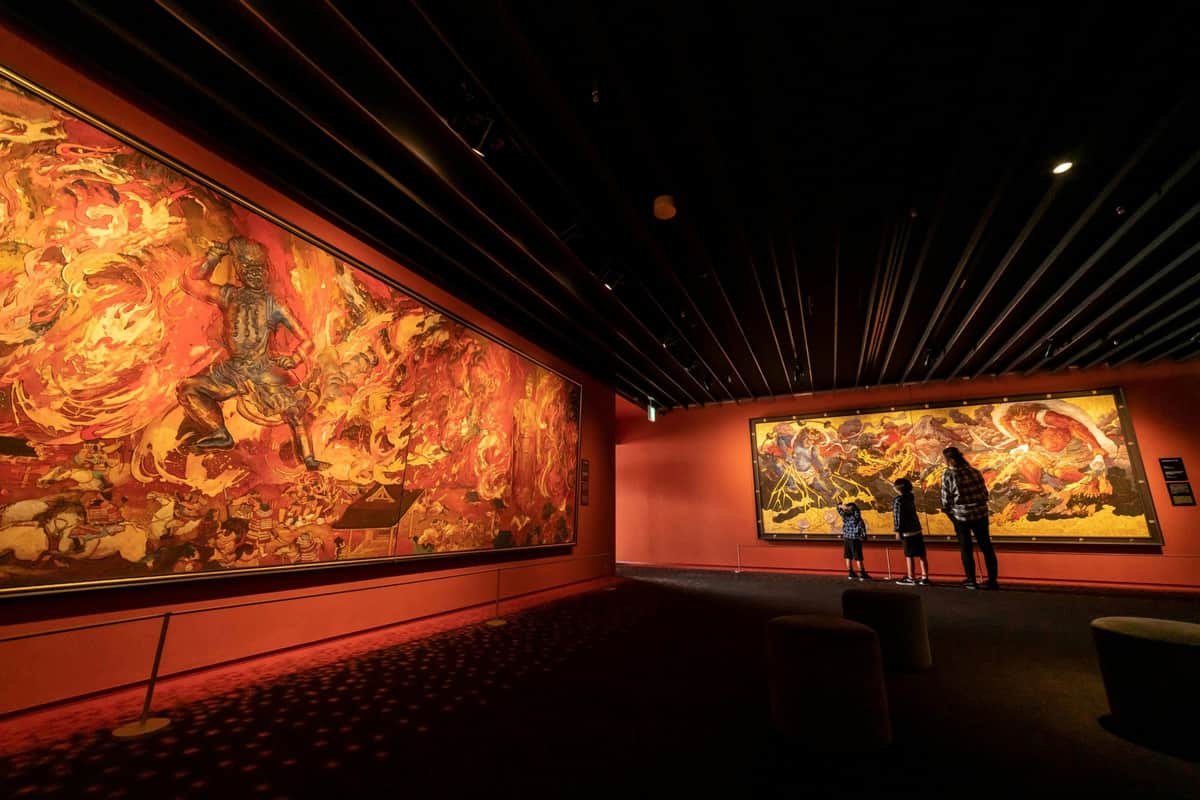 絹谷浩二天空美術館大阪:入場券+スカイガーデンへのアクセス 薄暗いアートギャラリーで、壁には大きくて鮮やかな絵画が描かれていて、それを見ているのは少人数のグループです。
