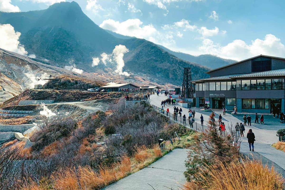 Mount Fuji & Hakone: Guided Day Tour from Tokyo + One-way Bullet Train Ticket A mountainous area with paths and buildings, people walking, and steam rising from vents, under a partly cloudy sky.