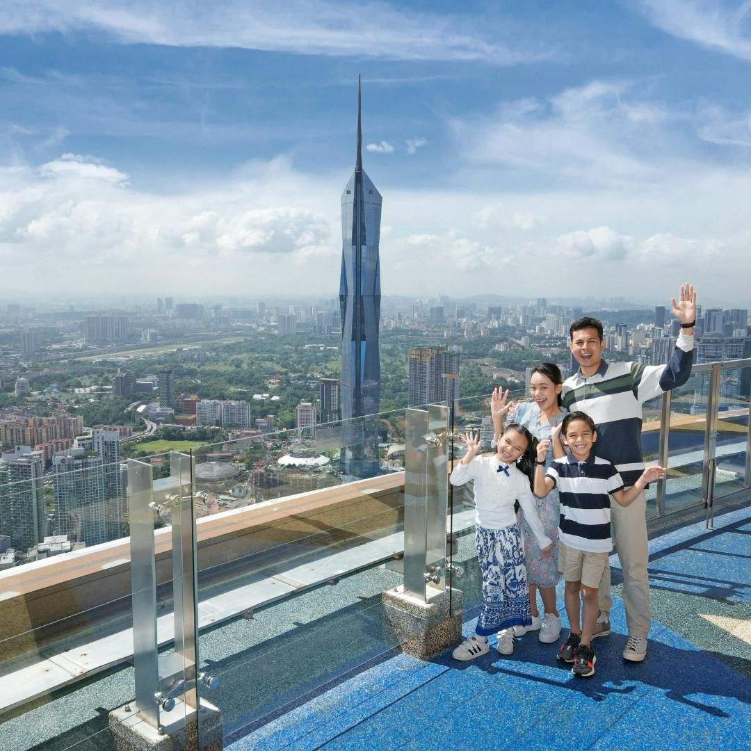 KL Tower: Entry Ticket A family of four waves from an observation deck with a cityscape and a tall, pointed skyscraper in the background.