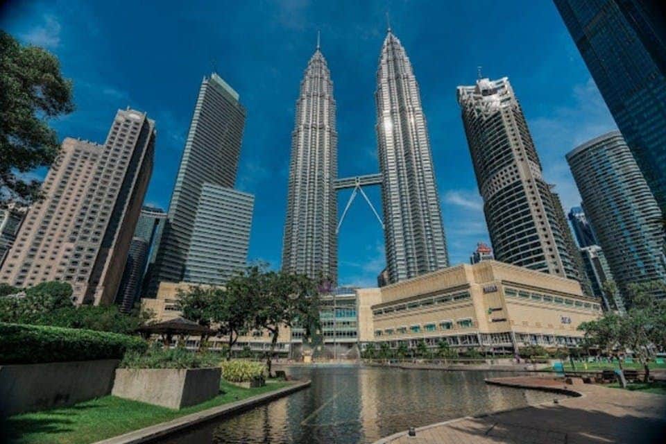 Petronas Twin Towers: Entry Ticket A view of the Petronas Twin Towers surrounded by other buildings, with a park and water feature in the foreground.