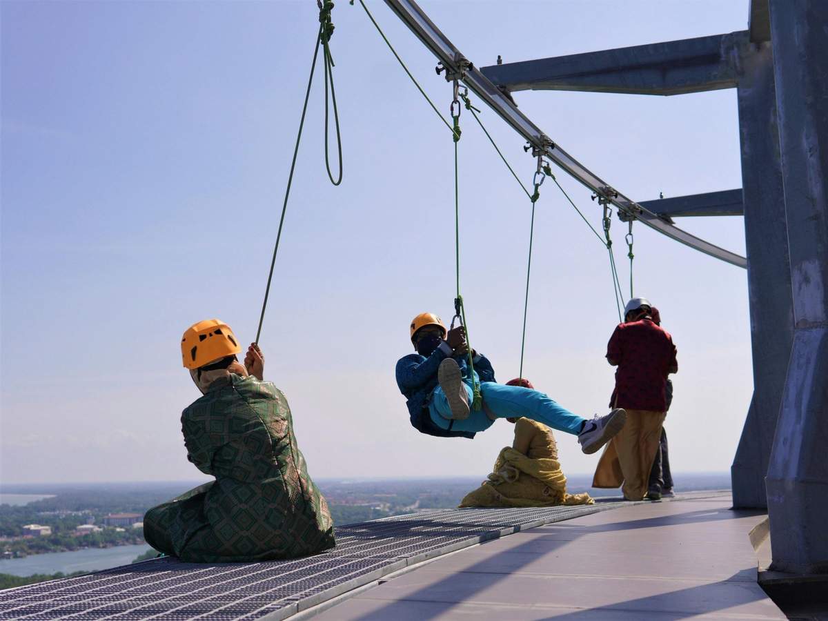 Kuantan 188 Tower: Skywalk-Erlebnis Menschen mit Sicherheitsausrüstung genießen eine Aktivität in großer Höhe, während sie an Klettergurten und Seilen vor dem Hintergrund eines klaren Himmels schweben.