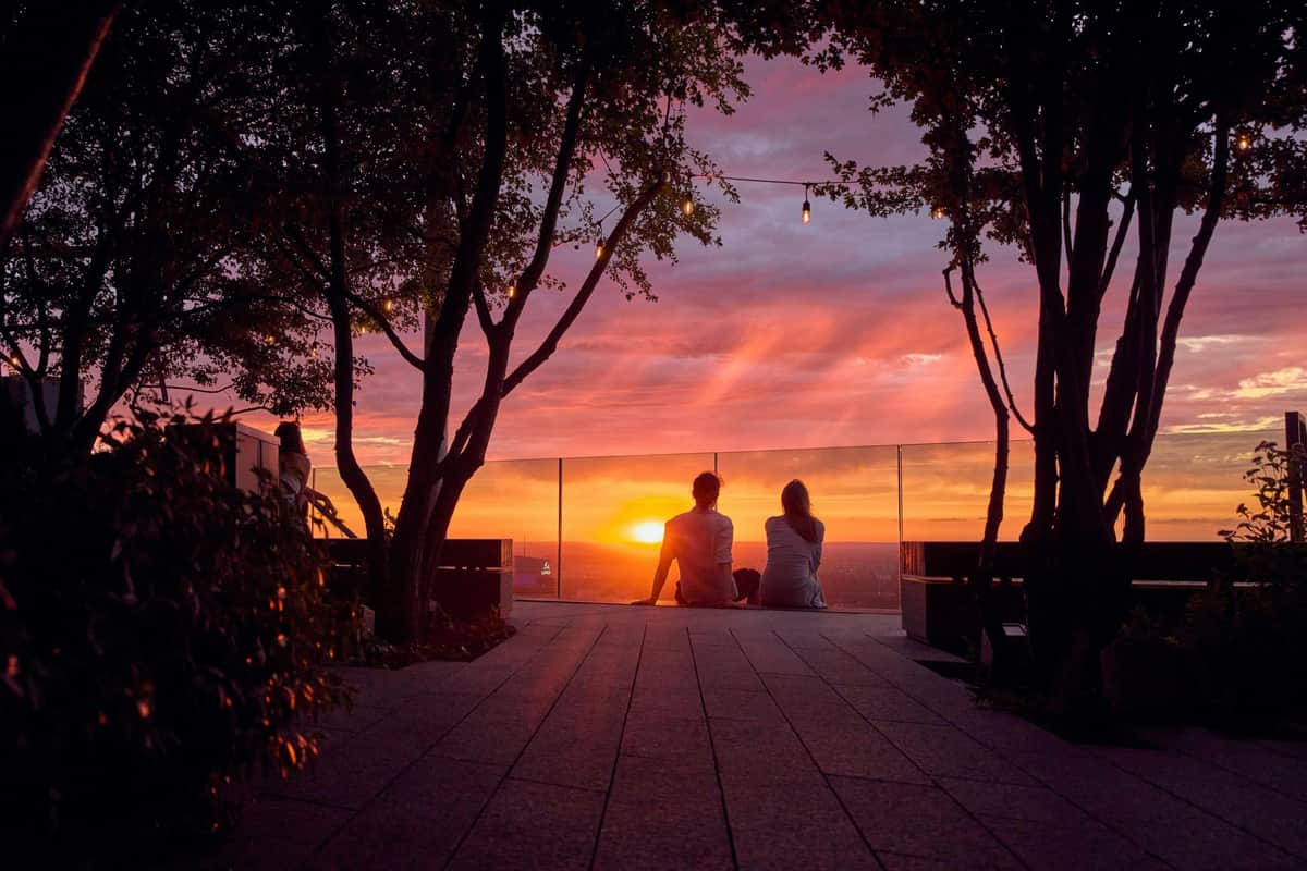Highline Warsaw: Entry Ticket + Rooftop Bar Access Two people sit on a rooftop floor, watching a vivid sunset through glass barriers, surrounded by trees and string lights.