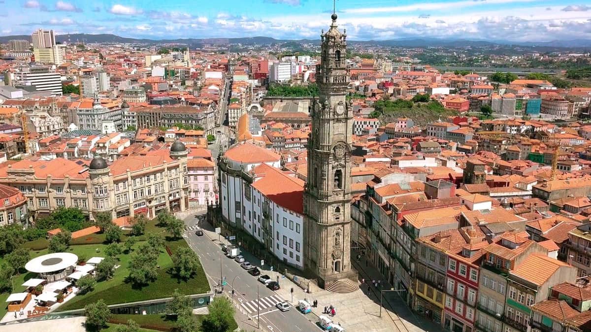An aerial view of a cityscape with a tall clock tower, red-roofed buildings, and a tree-lined park.
