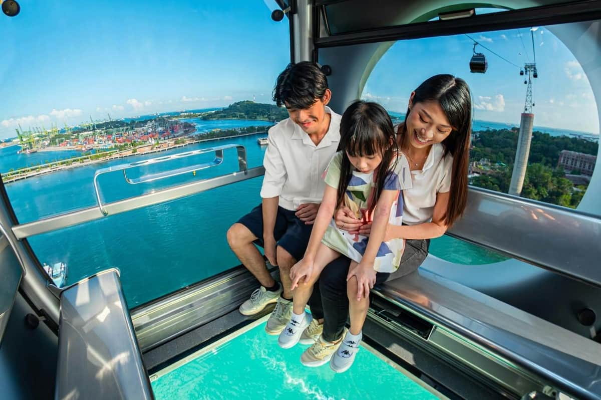 Teleférico de Singapur: SkyPass Premium Una familia se sienta en el interior de un teleférico con suelo de cristal, disfrutando de una vista panorámica del agua y del paisaje circundante.