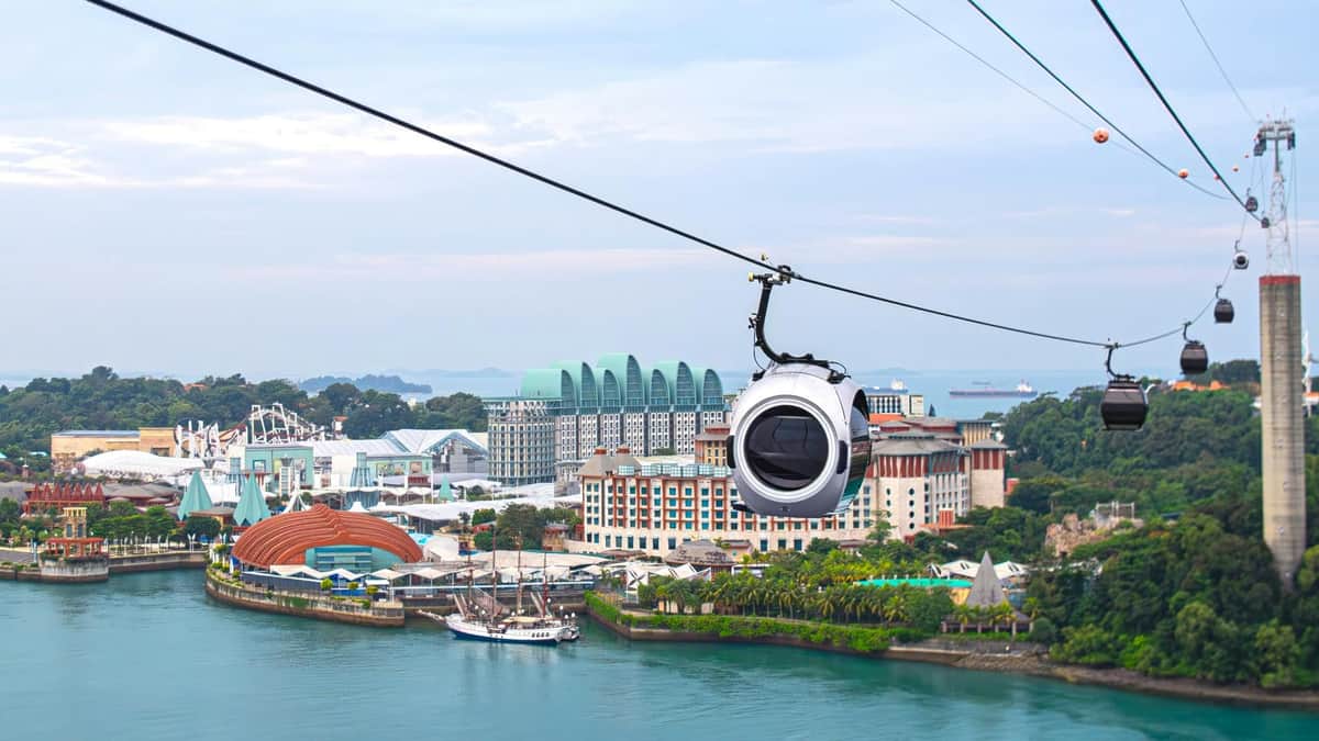 SkyPass del teleférico de Singapur: billete de ida y vuelta + comida infantil Arbora Un teleférico recorre una zona costera con edificios modernos y zonas verdes, con un cielo nublado de fondo.