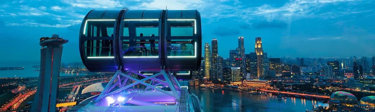 Singapore Flyer + Time Capsule Deux personnes à l'intérieur d'une nacelle d'observation éclairée sur une grande roue surplombent la ligne d'horizon d'une ville et le front de mer au crépuscule.