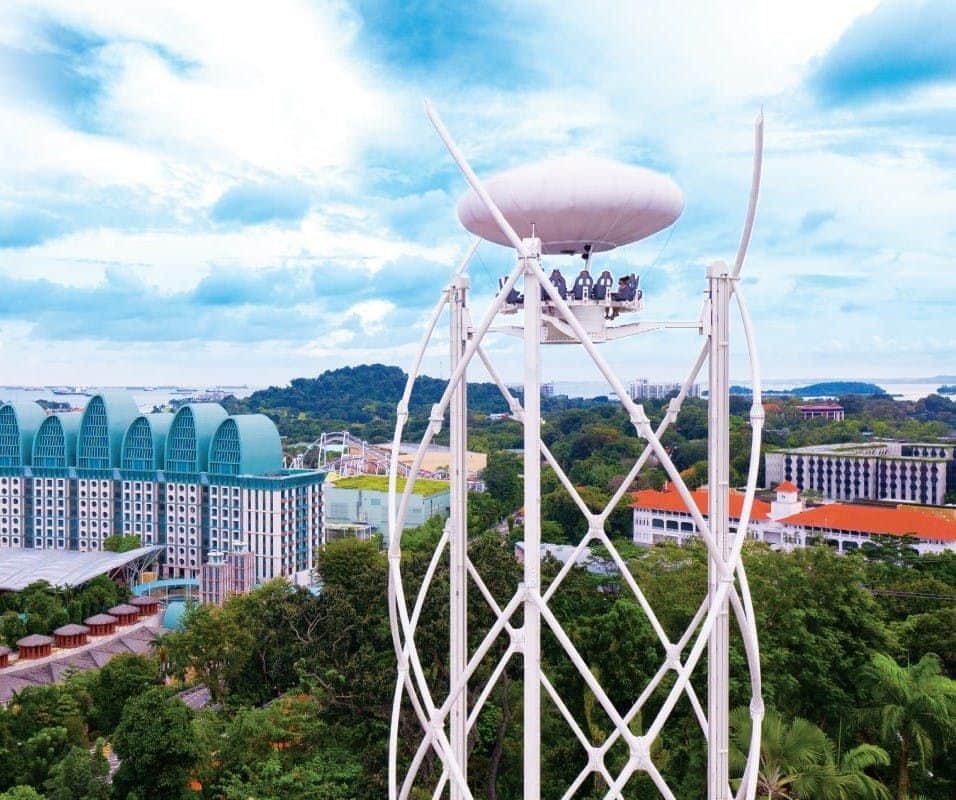 SkyHelix Sentosa Blick auf eine hohe Aussichtsplattform mit Menschen, die oben sitzen, umgeben von Gebäuden und üppigem Grün unter einem teilweise bewölkten Himmel.