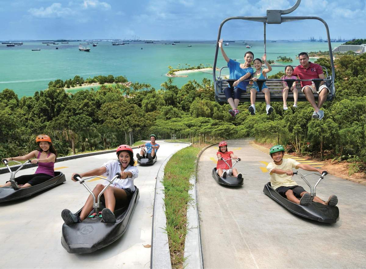 Skyline Luge Sentosa: Eintrittskarte Menschen, die mit Schlitten eine Bahn hinunterfahren, während andere auf einem Sessellift winken; üppiges Grün und ein malerischer Blick auf das Meer im Hintergrund.