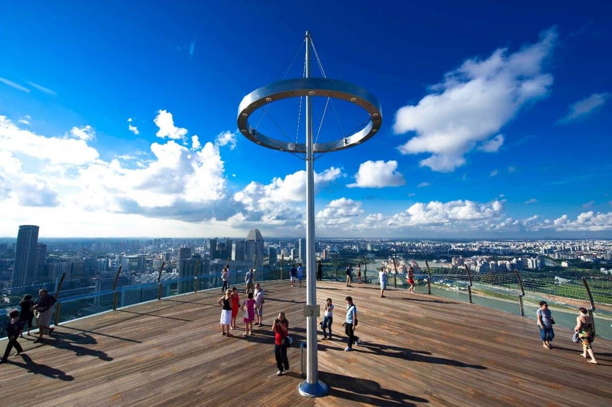 Entradas para la Cubierta de Observación del SkyPark: Entrada de un día La gente está de pie en una cubierta de madera con una alta estructura metálica, con vistas al perfil de la ciudad y al río, bajo un cielo azul brillante con nubes.