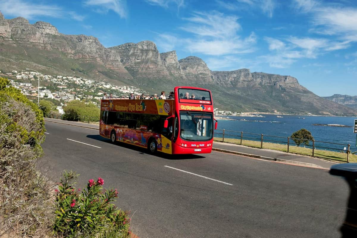 Red double-decker tour bus on a coastal road with mountainous terrain and a bay in the background under a clear blue sky.