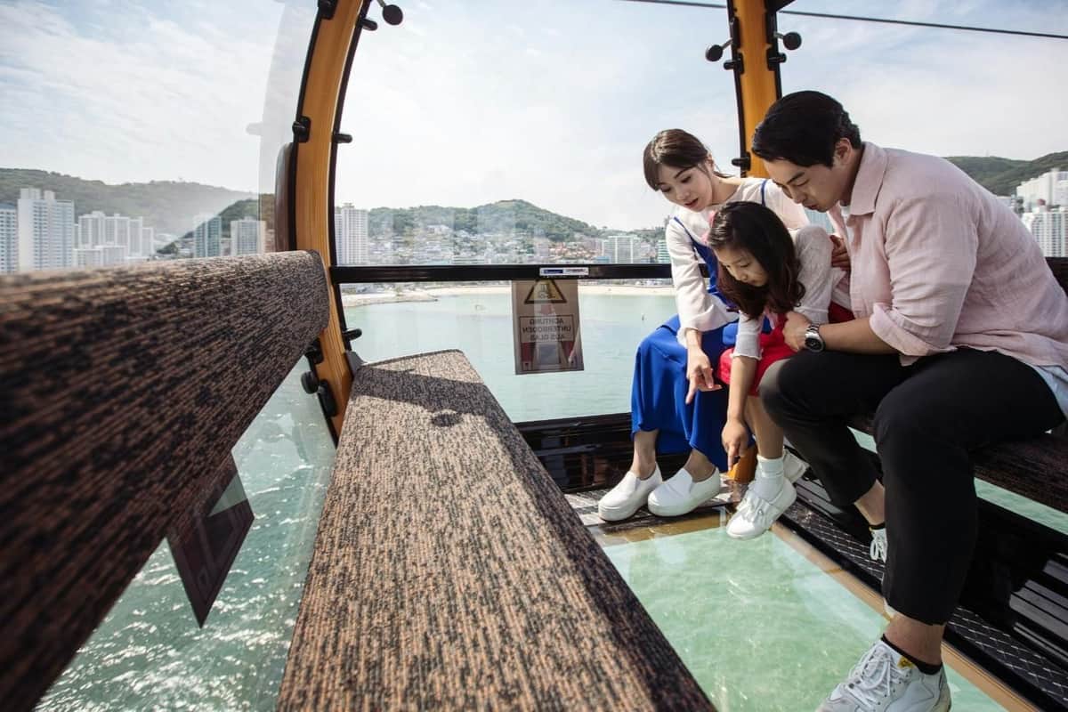 Songdo-kabelbaan: retourtrip – De kabelbaan van Songdo (Busan) Drie mensen zitten in een gondel met een glazen bodem en kijken door het glas naar het landschap beneden. Buiten zie je een stadsgezicht.