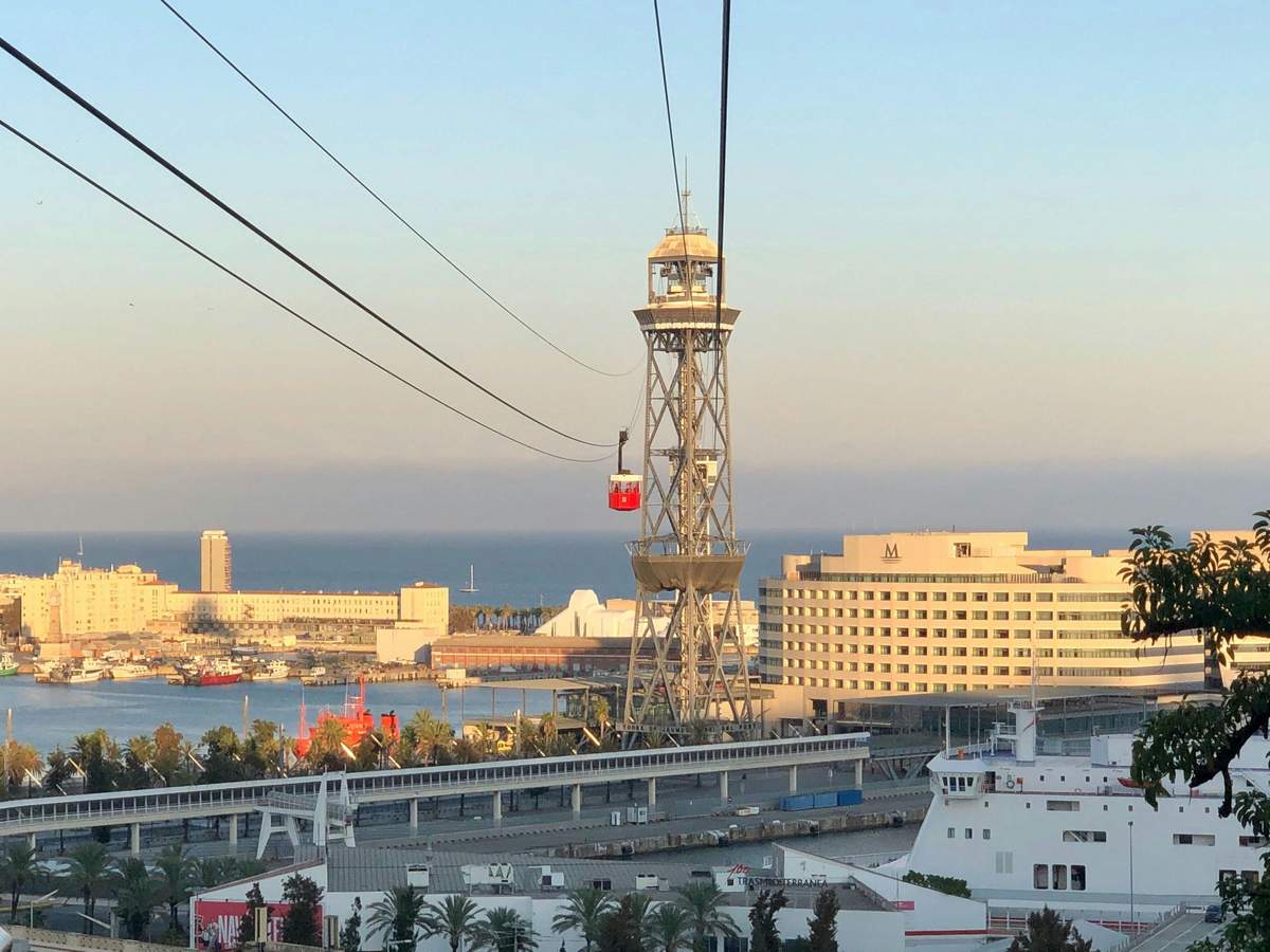 Barcelona Cable Car: Roundtrip from Barceloneta Beach A red cable car travels over a coastal cityscape with a tower, buildings, palm trees, and a harbor in the background.