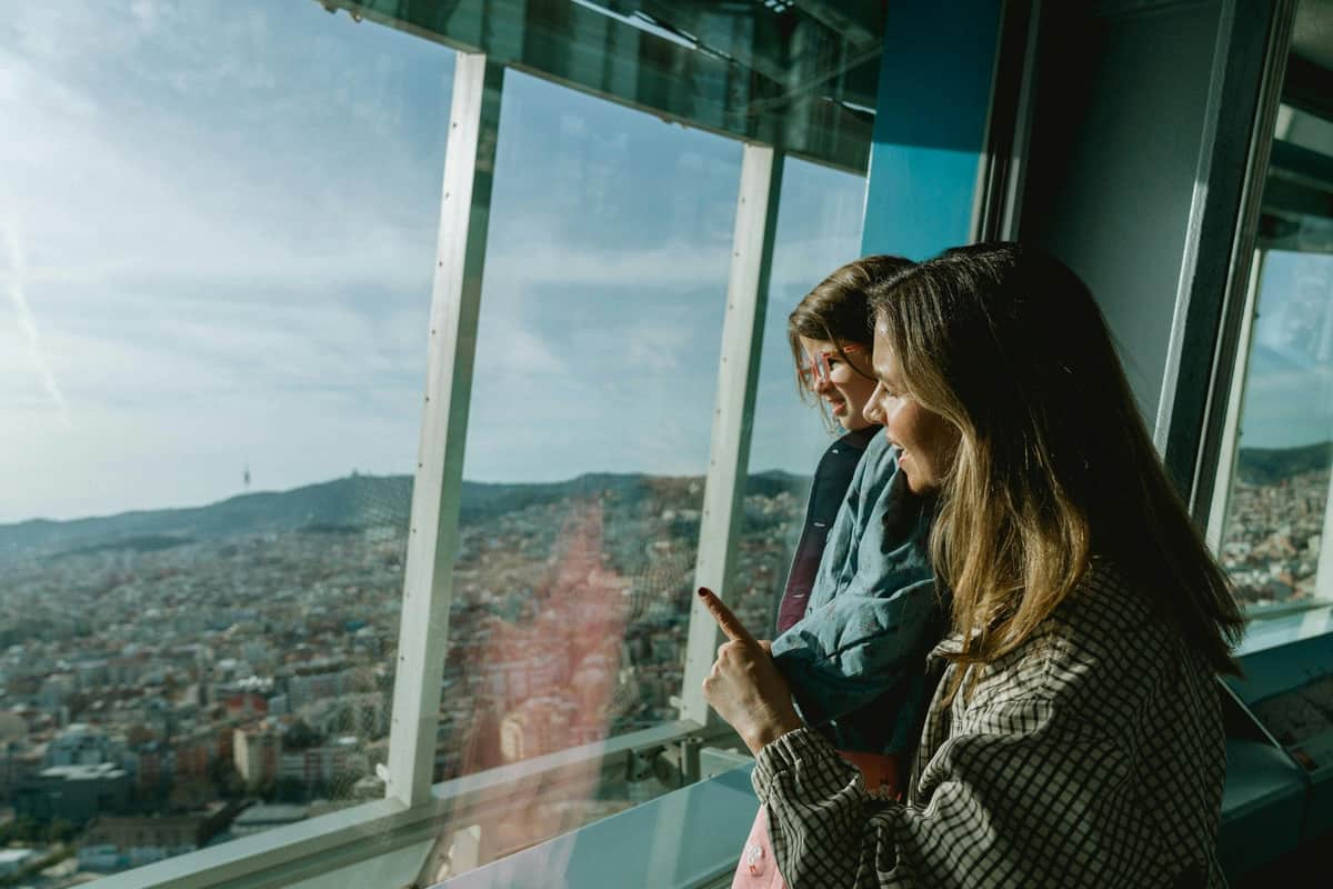 Mirador Torre Glòries: Open Date Ticket + Digital Audio Guide Two people are standing indoors near a large window, looking out at a panoramic view of the city beneath a bright, clear sky.