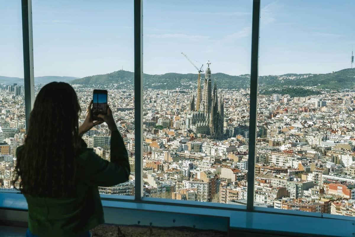 Mirador Torre Glòries: Skip The Line Ticket A person photographing the cityscape of Barcelona through a large window, with the Sagrada Família visible in the distance.