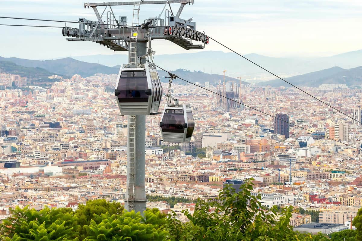 Montjuïc Cable Car: Roundtrip Ticket Two cable cars travel over a densely built city with mountains in the background and greenery in the foreground.