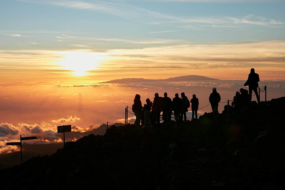 Mount Teide: Sunset Tour, Cable Car + Picnic Silhouette of people standing on a rocky path at sunrise, overlooking a sea of clouds and distant mountains in the backdrop.