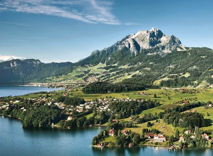 Mount Pilatus with Transport from Zurich Scenic landscape of a lakeside village with green fields, scattered houses, and a large mountain range in the background under a blue sky.
