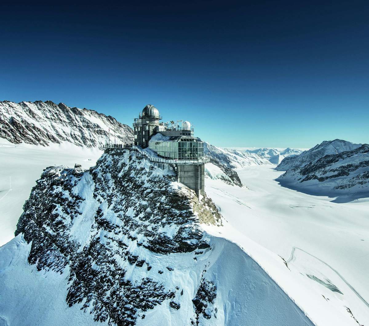 Jungfraujoch with Transport from Zurich Observatory with dome perched on a snow-covered mountain peak, surrounded by vast snowy landscape and clear blue sky.