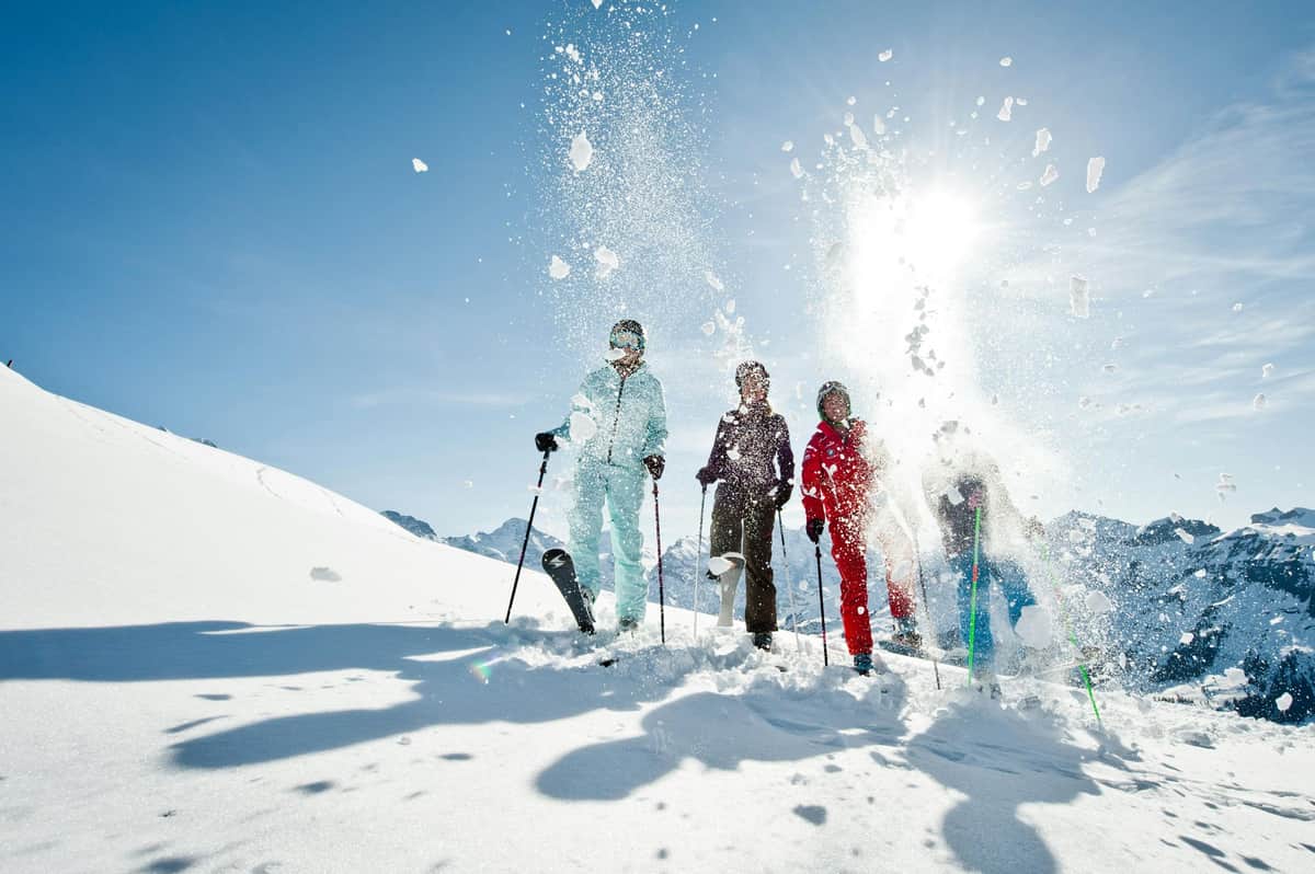 Swiss Ski Experience at Jungfraujoch with Transport from Zurich Four skiers in colorful outfits stand on a snowy slope, with snow flying around them and bright sunlight in the background.