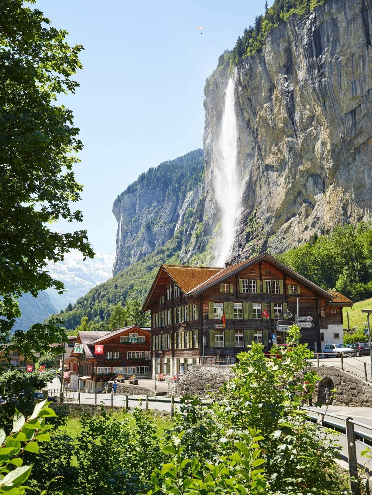 Grindelwald, Interlaken & Lauterbrunnen: Day Trip + Transport Rustic buildings with red roofs near a tall waterfall cascading down a cliff, surrounded by lush greenery and mountains.