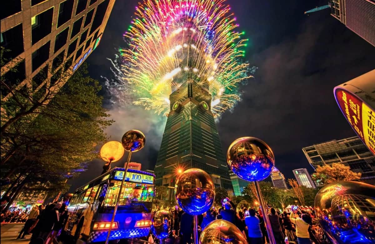 Taipei 101 Observatory Colorful fireworks explode above a tall building at night, with a crowd below and reflective spheres in the foreground.